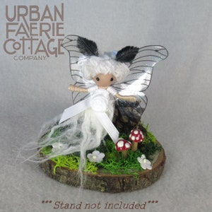 May include: White fairy doll with black wings and white yarn hair, standing on a wooden base with moss and mushrooms.  The doll is wearing a white dress and a white ribbon.  ***Stand not included***