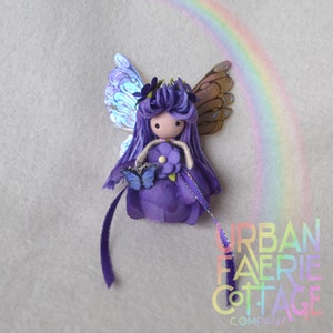 May include: Purple fairy doll with iridescent wings, holding a purple flower and a blue butterfly charm. The doll has long purple yarn hair and a flower crown. The background is a light grey with a rainbow.