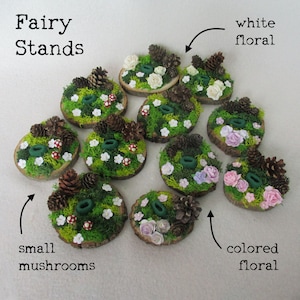 May include: Fairy stands with a variety of floral arrangements. Some stands feature white flowers, while others have pink, purple, and white flowers. All stands are decorated with moss, small mushrooms, and pine cones.