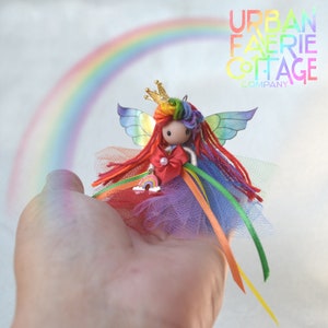 May include: Rainbow fairy doll with a crown, wings, and a rainbow-coloured dress. The doll is holding a rainbow charm. The doll is in front of a rainbow background.