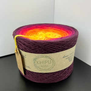 May include: A skein of yarn with a gradient of colors from dark purple to yellow. The yarn is wound around a cardboard tube with a label that says "KHIPU" and "Anna Sock Yarn".