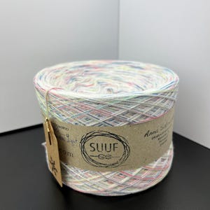 May include: A skein of white yarn with multi-coloured flecks. The yarn is wrapped around a cardboard tube with a brown label that says "SUUF" and "Anna Sol Crochet".