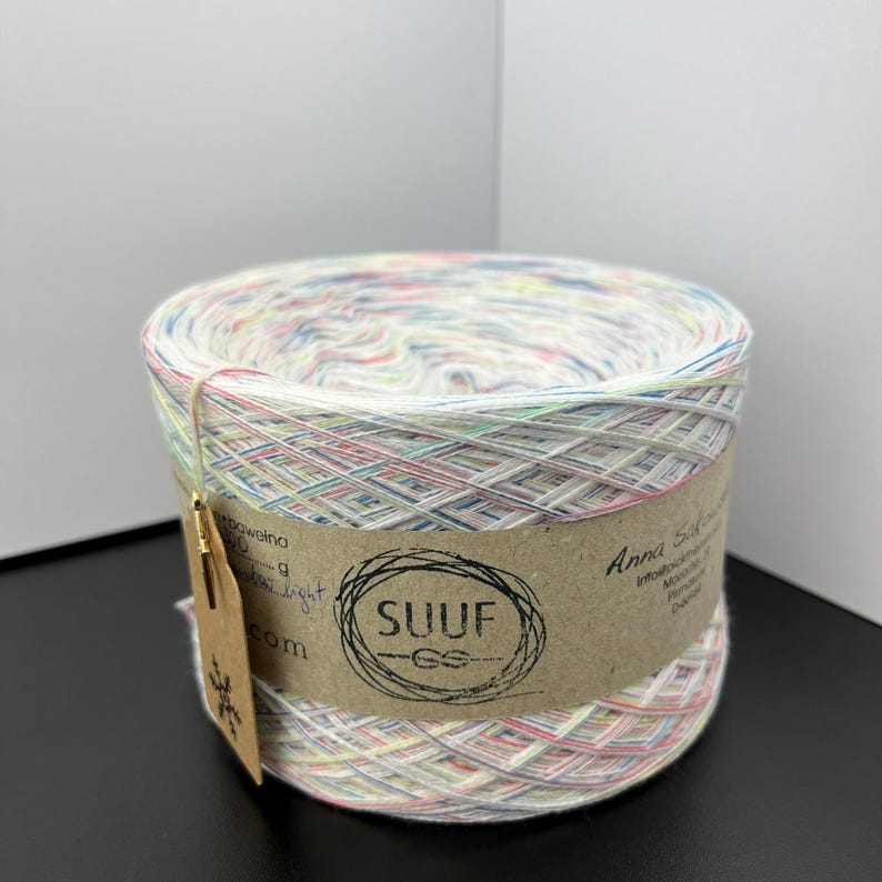 May include: A skein of white yarn with multi-coloured flecks. The yarn is wrapped around a cardboard tube with a brown label that says "SUUF" and "Anna Sol Crochet".