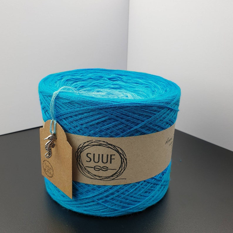 May include: A skein of turquoise blue yarn with a white ombre effect. The yarn is wrapped around a cardboard tube with a brown paper label that says "SUUF" with a knot symbol. A silver seahorse charm is attached to the label.