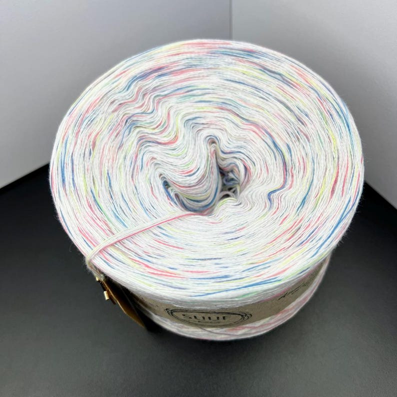 May include: A white ball of yarn with multi-coloured speckles of pink, blue, green, and yellow. The yarn is wound around a cardboard tube with a label that says "SHUF".