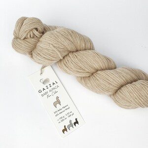 May include: A skein of beige yarn labelled "GAZZAL BABY ALPACA Pure Color" with the composition 55% Baby Alpaca and 45% Merino Wool. The yarn is 100 grams (3.52 ounces) and 320 metres (349 yards) long.