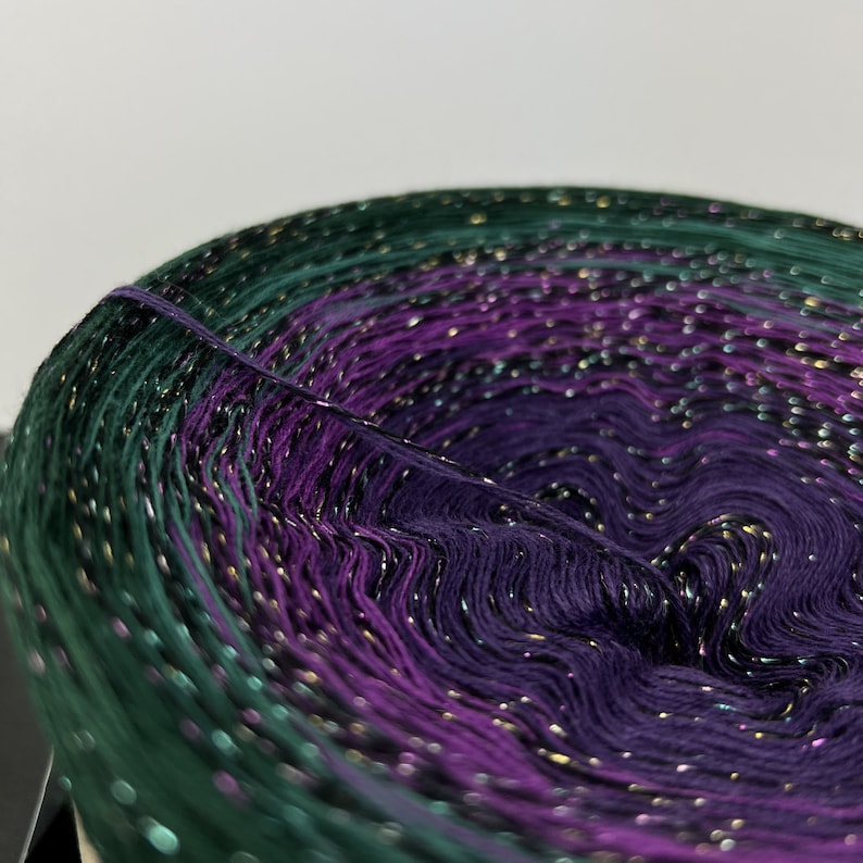 May include: A skein of yarn with a gradient of green, purple, and black colors. The yarn has a metallic gold thread woven throughout.