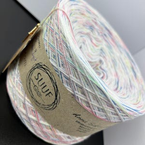 May include: A skein of white yarn with multi-coloured stripes. The yarn is wound around a cardboard tube with a brown label that says "SUUF" and "Anna Salo".