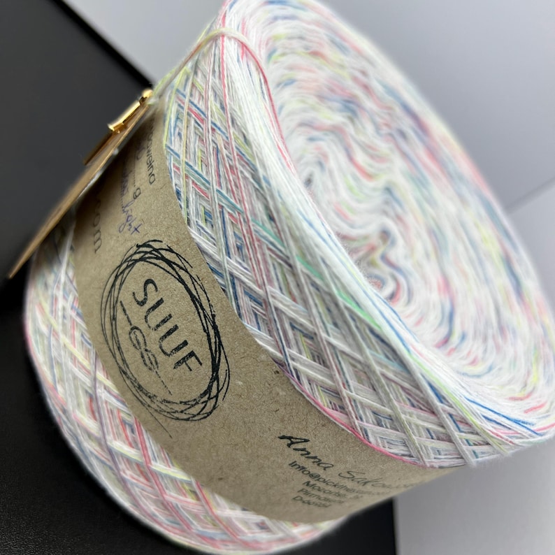 May include: A skein of white yarn with multi-coloured stripes. The yarn is wound around a cardboard tube with a brown label that says "SUUF" and "Anna Salo".