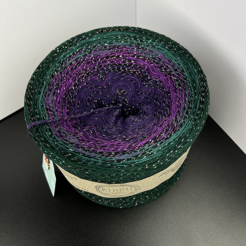 May include: A skein of green, purple, and gold yarn with glitter. The yarn is wound into a ball and has a label that says "KHIPU".