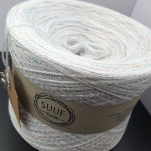 May include: A white ball of yarn with silver, gold, and pink glitter threads. The yarn is wrapped around a brown label with the word "SUUF" printed on it.