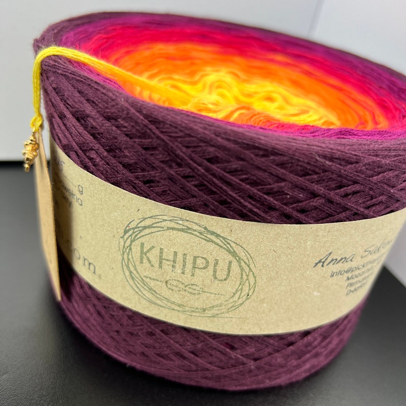 May include: A skein of yarn in a gradient of burgundy, orange, and yellow colors. The yarn is wound around a cardboard tube with a brown paper label that reads "KHIPU" in a green circle. The label also includes the text "Anna Saavedra" and other text that is too small to read.