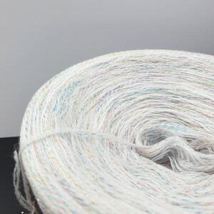 May include: A close-up of a white yarn with iridescent glitter threads woven throughout. The yarn is wound into a ball shape.