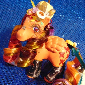 Puede incluir: Un juguete de pony pintado a medida, colorido, con una melena y cola arcoíris. El pony lleva una corona con cristales y otros elementos decorativos. El pony está de pie sobre una superficie azul brillante.