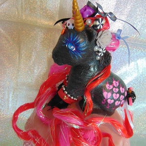 May include: A black unicorn figurine with a gold horn, adorned with a skull, pearls, and a purple accent. The unicorn has red and pink hair, blue eye makeup, and a broken heart design. It sits atop a pink, scalloped base.