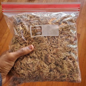 May include: A clear plastic bag with a red zipper closure, filled with dried, light brown moss. The bag has a label with spaces for "CONTENTS:" and "DATE:" and is held by a hand.