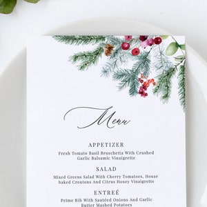 Puede incluir: Una tarjeta de menú blanca con un diseño de acuarela de ramas de pino, bayas rojas y piñas. El menú enumera opciones de aperitivo, ensalada, plato principal y postre. El menú está en un plato blanco con vegetación y una vela en el fondo.