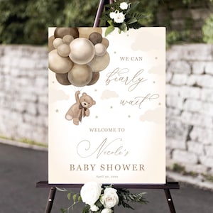 Pode incluir: Uma placa de chá de bebé num cavalete com um ursinho a segurar balões. A placa diz "We can nearly wait" e "Welcome to Nicole's Baby Shower". A placa é decorada com flores e tem uma paleta de cores neutras.