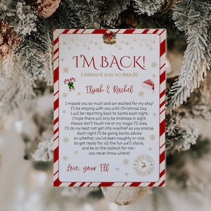 Puede incluir: Una tarjeta navideña con un borde a rayas rojas y blancas, colgada en un árbol de Navidad. La tarjeta dice "I'M BACK!" e incluye un mensaje de "Elijah & Rachel" con ilustraciones festivas.