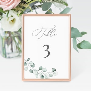 May include: A white table number card with a gold frame and a green eucalyptus leaf design. The card says "Table 3" in a cursive font.