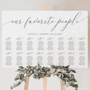 May include: A white wedding seating chart with the text "our favourite people" in cursive script. The chart lists the names of guests assigned to each table number. The chart is on a wooden easel with greenery.