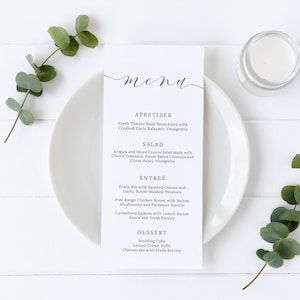 May include: A white menu card with a handwritten 'menu' title and a list of appetizers, salads, entrees, and desserts. The menu is on a white plate with a sprig of eucalyptus on the left and a candle on the right.