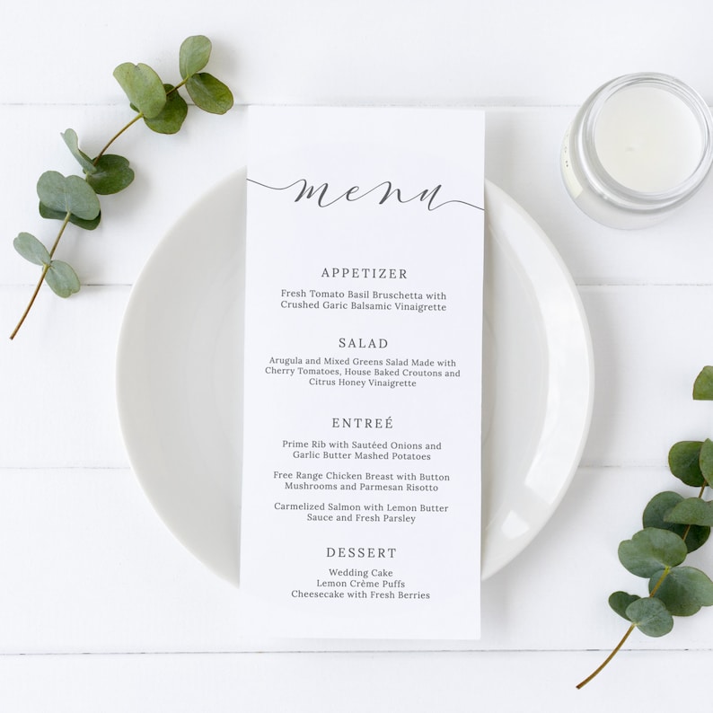 May include: A white menu card with a handwritten 'menu' title and a list of appetizers, salads, entrees, and desserts. The menu is on a white plate with a sprig of eucalyptus on the left and a candle on the right.