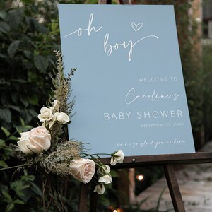 Pode incluir: Uma placa de boas-vindas azul claro com caligrafia branca que diz "Oh boy" com um pequeno cora&ccedil;&atilde;o acima da palavra "boy". A placa tamb&eacute;m inclui o texto "Bem-vindo ao Chuveiro de Beb&ecirc; da Caroline, 23 de setembro de 20XX, estamos muito felizes que esteja aqui".