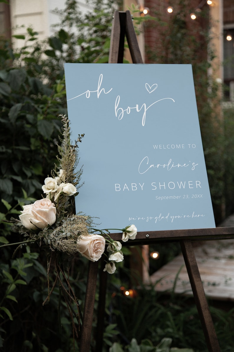 Pode incluir: Uma placa de boas-vindas azul claro com caligrafia branca que diz "Oh boy" com um pequeno cora&ccedil;&atilde;o acima da palavra "boy". A placa tamb&eacute;m inclui o texto "Bem-vindo ao Chuveiro de Beb&ecirc; da Caroline, 23 de setembro de 20XX, estamos muito felizes que esteja aqui".