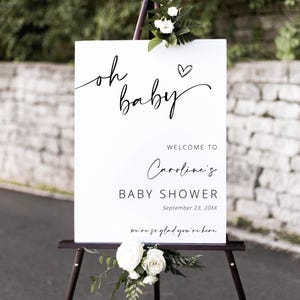 May include: A white sign on a dark brown easel, decorated with white flowers and greenery. The sign reads "oh baby" with a heart, "Welcome to Caroline's Baby Shower" and the date. The sign is set outdoors.
