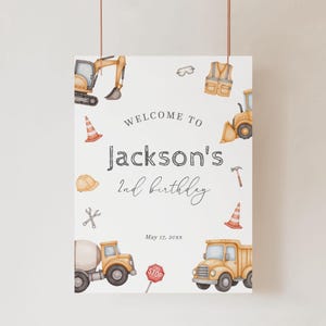 May include: A personalized birthday party sign with construction-themed illustrations. The sign reads "Welcome to Jackson's 2nd birthday" with construction vehicles, safety gear, and the date May 17, 20xx.