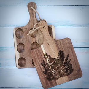 May include: Two wooden cutting boards, one with a design of a hen and flowers, the other with six egg-shaped indentations.