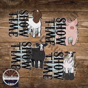 May include: A digital graphic featuring a goat, pig, cow, and sheep with the words "Show Time" in a distressed font. The animals are in a cartoon style, set against a wood grain background. A logo is in the bottom left corner.