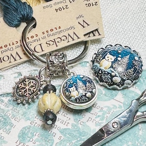May include: A silver key chain with a snowflake charm, a white bead, and two glass owl charms. The owl charms are round and feature a blue background with white owls and a white snowflake. The key chain is on a white fabric background with text that reads "Weeks D Works" and "Specializing in Hand Dyed Fibers".