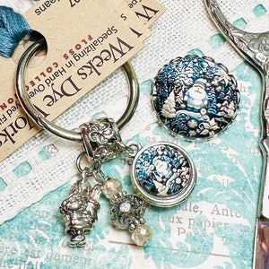 May include: A silver key chain with a round charm featuring a winter scene with a snowman. The charm is attached to a silver ring with a smaller charm of a snowman and a few crystal beads. The key chain is on a white background with a blue and white patterned fabric.