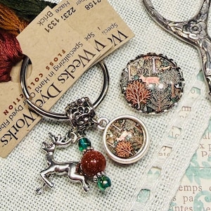 May include: An assortment of craft supplies including embroidery floss, a deer charm, decorative buttons, and vintage scissors. The floss is in shades of green, orange, and red. The buttons feature a forest scene with a deer.