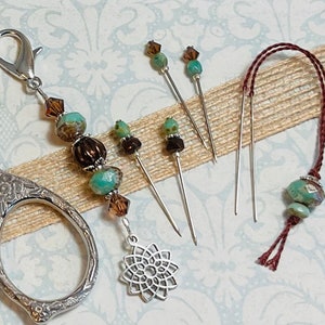 May include: A set of sewing tools with a beaded needle minder, a pair of scissors, and four sewing pins with turquoise and brown beads. The tools are on a beige fabric background.