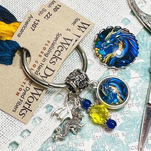 May include: A silver key chain with two blue and gold dragon charm pendants, a silver dragon charm, and yellow and blue glass beads. The key chain is on a white fabric background with a tag that reads "W Weeks Dy Works" and "Specializing in Hand Dyed Fibers".