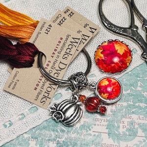 May include: A seasonal collection featuring a silver pumpkin charm keyring, leaf-patterned cabochons, embroidery floss, and scissors. The keyring has a pumpkin charm and red beads. The floss colours are orange and burgundy.