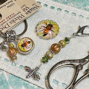 May include: A silver key chain with a bee charm and a glass bee charm. A silver scissor with a silver bead chain with green and brown beads and a bee charm.