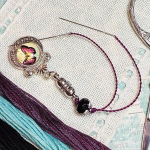 May include: A silver needle threader with a butterfly charm and a black bead. The needle threader is on a piece of white fabric with several strands of embroidery floss in black, teal, and purple.