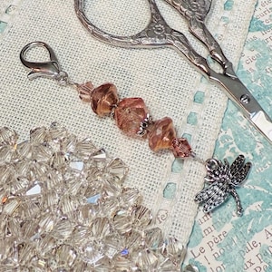 May include: A silver dragonfly charm with a beaded accent and a pair of ornate scissors. The charm features a cluster of amber-colored beads and a silver clasp. A collection of clear, faceted beads is also visible.