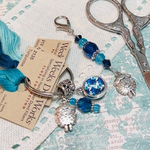 May include: An assortment of turquoise embroidery floss, a pair of silver scissors, and a charm with blue beads and a sheep pendant. The floss is labeled "Turquoise" and "Weeks Dye Works". The background has a floral pattern.