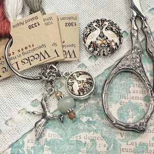 May include: A silver metal ring holding three skeins of embroidery floss in red, white, and olive green, with attached charms including a silver bird, a small bead, and a circular pendant featuring a white crane illustration on a black background.