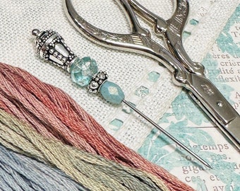 May include: A close-up of embroidery floss in shades of pink, green, blue, and purple. A silver needle with a decorative bead is lying on top of the floss. A pair of silver embroidery scissors are also in the image.