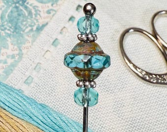 May include: A decorative pin with a teal and silver beaded design. The pin is next to teal and tan embroidery floss, and silver scissors. The background includes a vintage paper with a teal floral design.