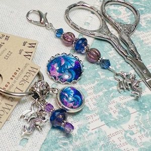 May include: A silver scissor charm with a blue and purple glass dragon charm, a silver dragon charm, and a silver key ring with a blue and purple glass dragon charm. The charm is attached to a tag that reads "Weeks De Works Specializing in Hanover Dyed Fibers FLOSS COLLECTION 2333 Pu Americana 23: 130 Peoria Purple Spe Spe"