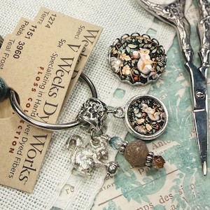 May include: A silver metal ring holding a collection of embroidery floss in shades of green and brown, a silver squirrel charm, a round pendant with a squirrel illustration, and a beaded dangle, alongside a pair of ornate silver scissors.