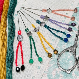 May include: A set of seven needle threaders with colorful beaded tassels. Each needle threader has a different charm, including a flower, a bee, a turtle, a seashell, a butterfly, and a heart. The threaders are arranged on a white fabric background with a pair of silver scissors.