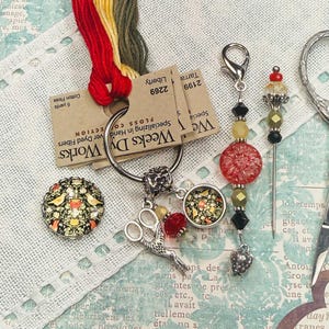 May include: A collection of sewing accessories including a round button with a bird and floral pattern, a metal ring with a small scissor charm and beads, a beaded needle minder, and silver-toned embroidery scissors with ornate handles.